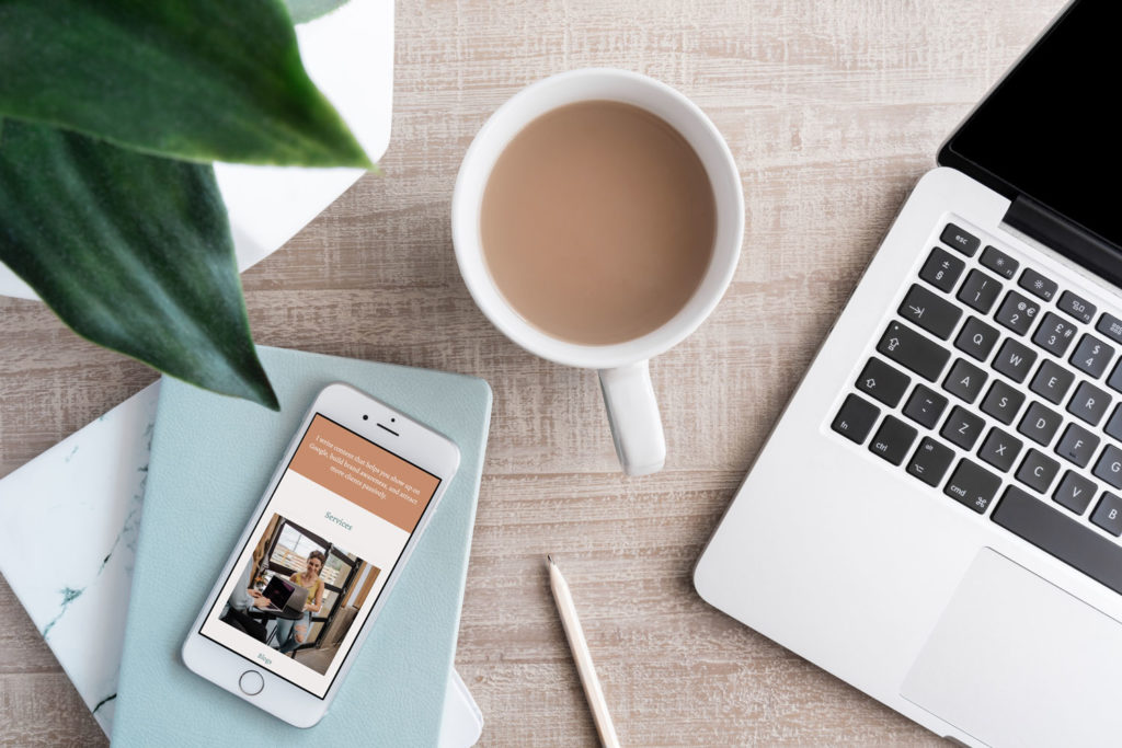 A Squarespace website is pulled up on a phone next to a mug and a laptop.