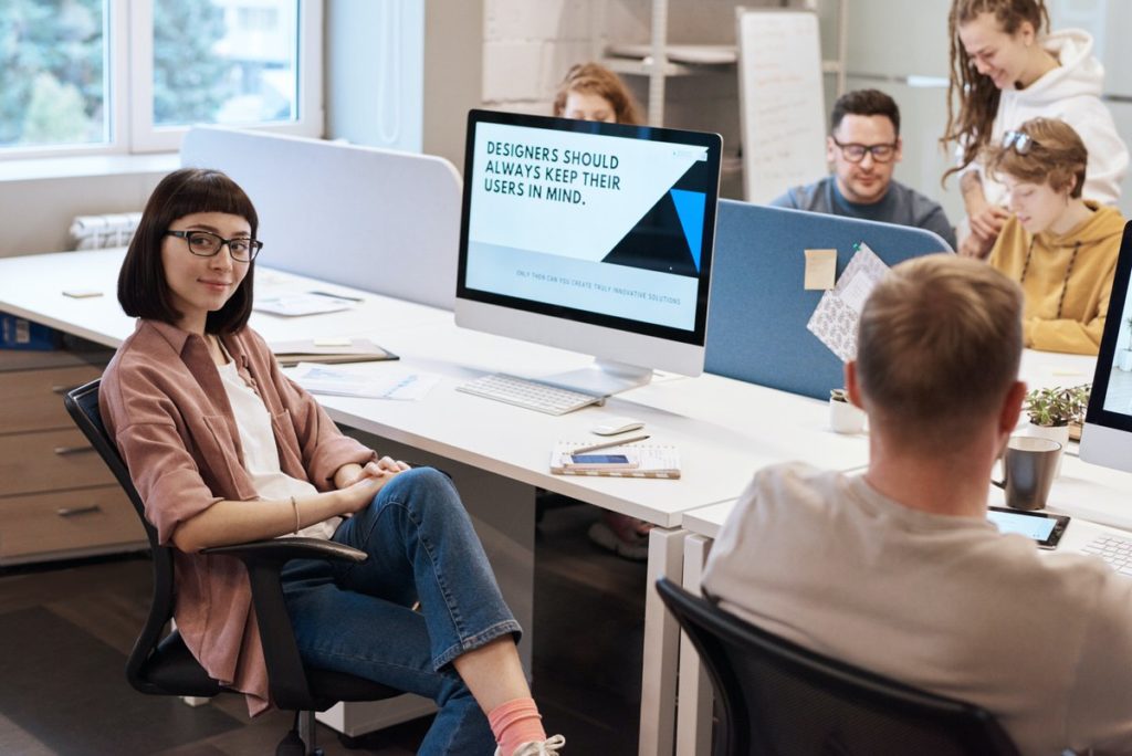 Girl sitting in a chair at a desk by coworkers smiling at the camera