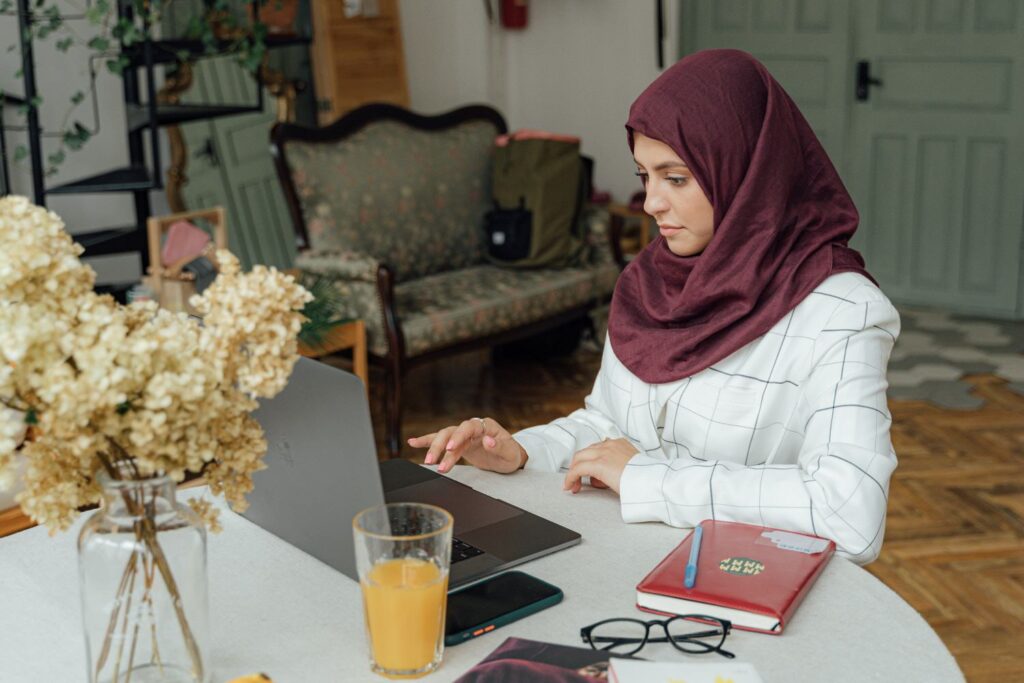 woman sitting at a round table working on a laptop with various notebooks and a drink next to her 
