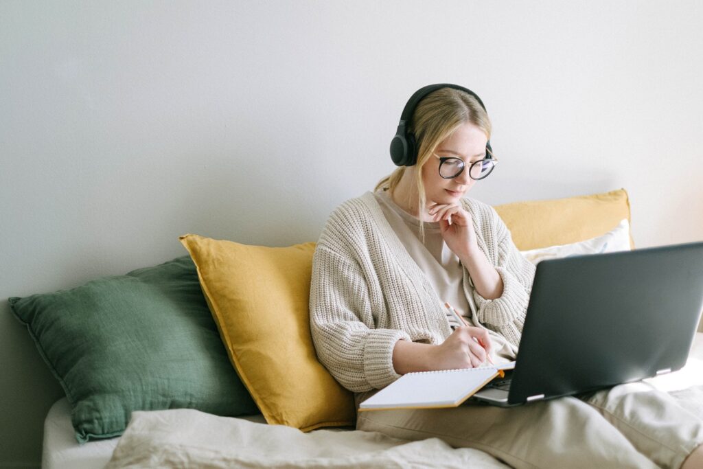 woman sitting in bed with headphones on writing in a notebook with her laptop open on her legs 