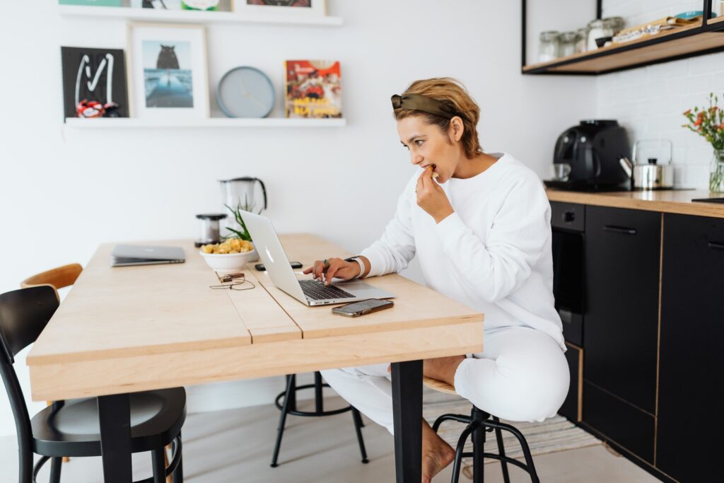 Woman sitting on a stool typing on her laptop while eating food 