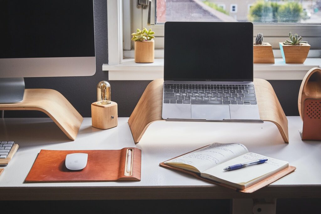 laptop and desktop on small stands on a desk with a notebook in front of them 