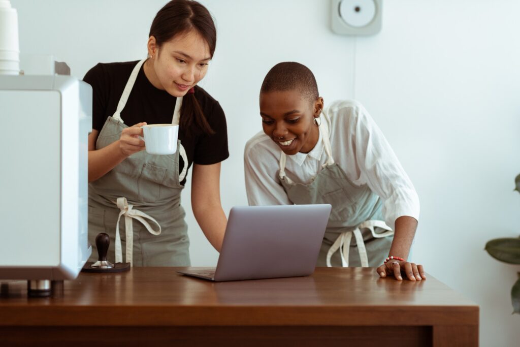 Two baristas at a coffee shop leaning on a counter looking at a laptop 