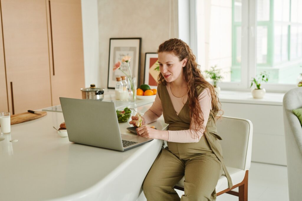 woman sitting at a kitchen island eating a sandwich as she works on her laptop 