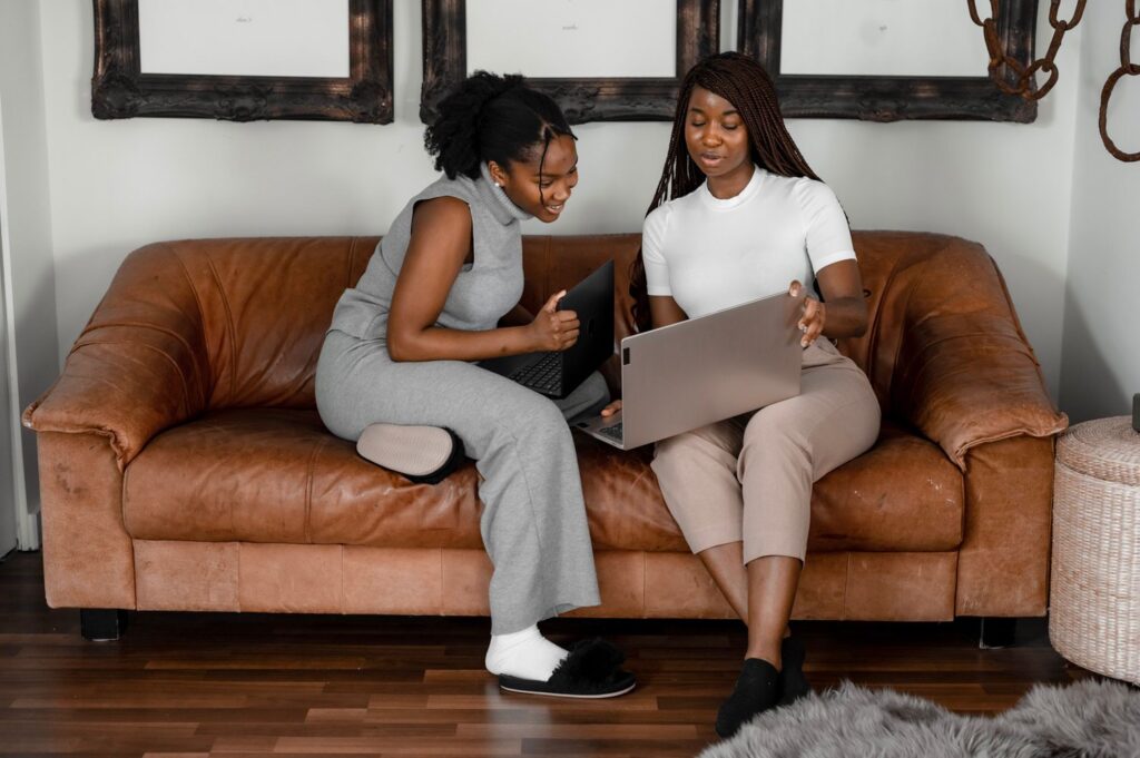 two women sitting on a couch together looking at a laptop 