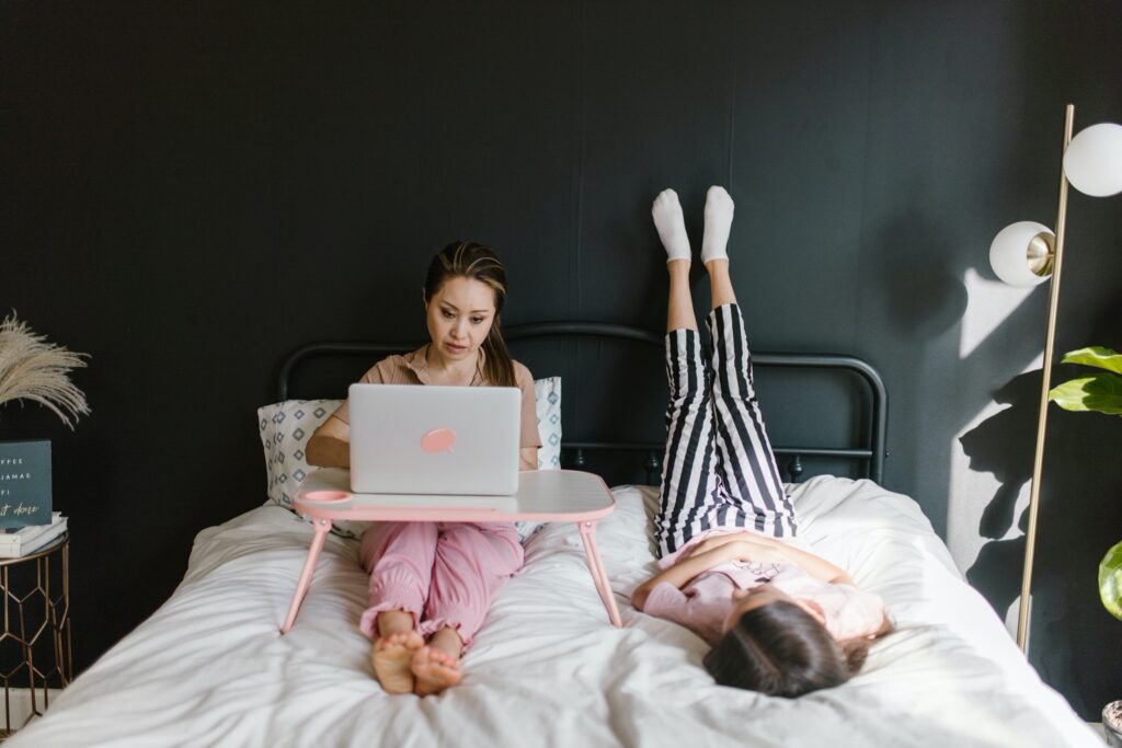 Two women laying in a bed. One is sitting up with her computer on a lap desk. The other is laying on her back with her feet propped up on the wall 