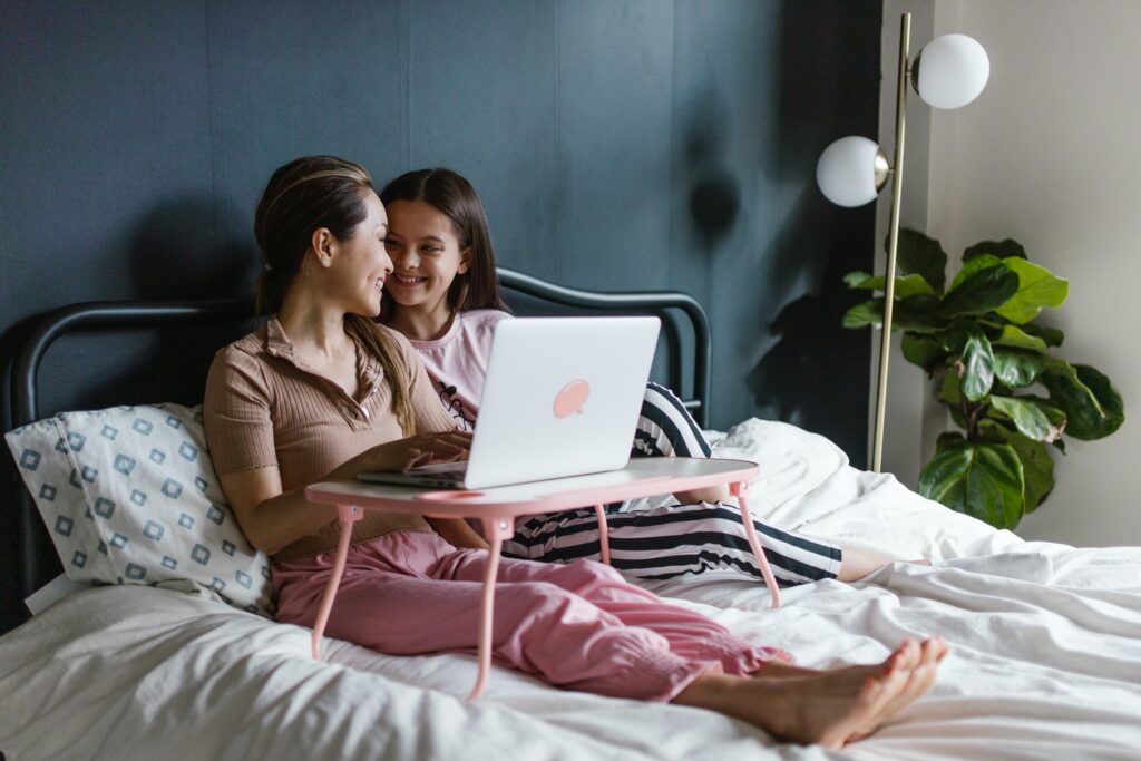 A mother and a daughter sitting in a bed as the mother works on a computer 
