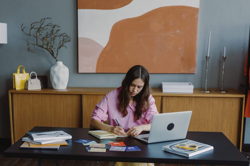 woman sitting at a long desk writing in a notebook with a laptop open in front of her
