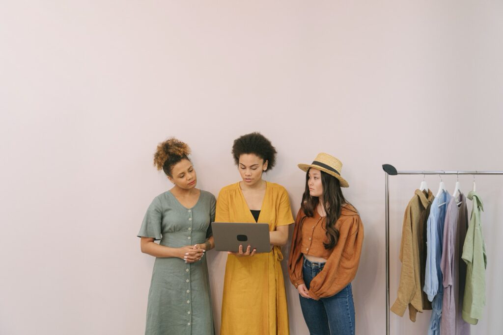 three woman standing against a wall looking at a laptop with a small clothing rack next to them 