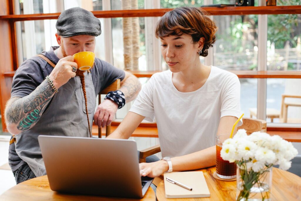 two people sitting at a small table at a cafe working on a computer as one sips coffee 
