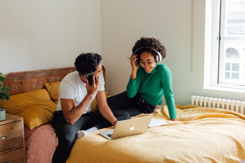 two people sitting on a bed looking at a laptop while one is on a phone call and the other is putting headphones on 