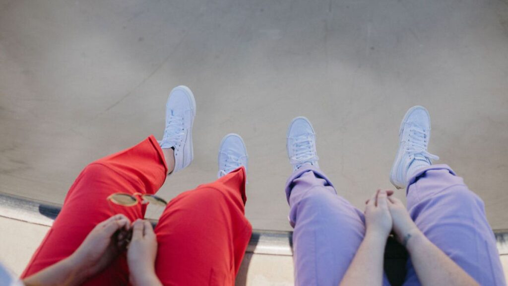 View looking down into a skateboard bowl with two people in red and purple pants sitting on the edge with their legs dangling into the bowl. 