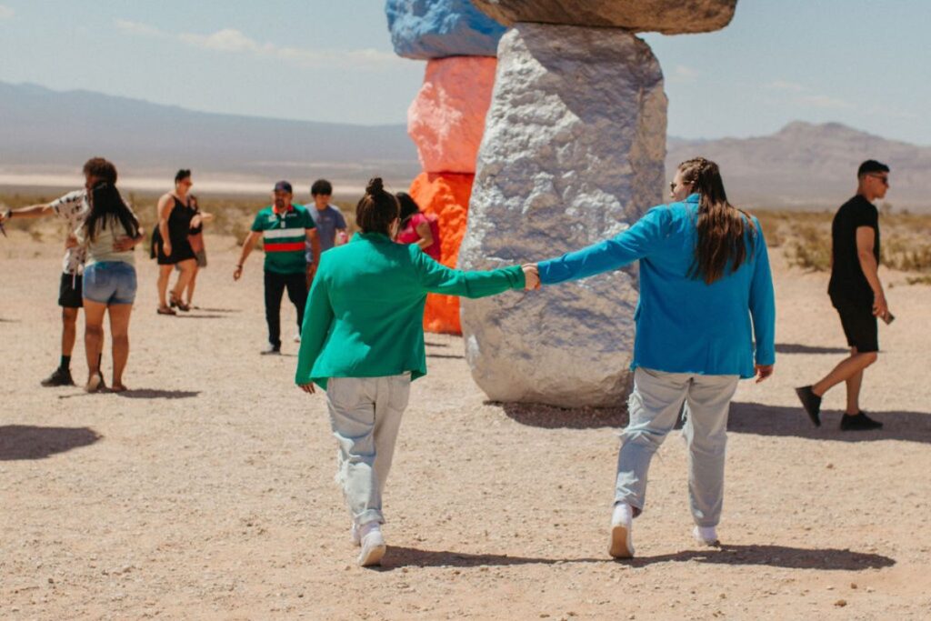 Two women holding hands as their arms are outstretched. They are walking in a desert area with colorful rocks and groups of people. 