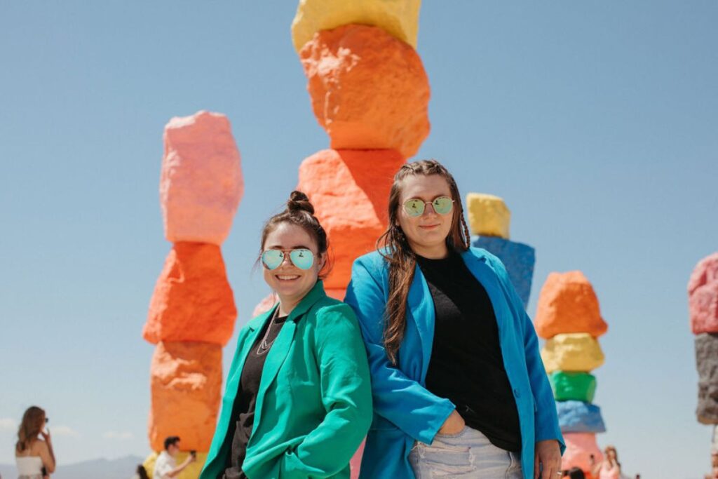 Two women in blazers and sunglasses leaning up against each other smiling with large stacks of colorful rocks behind them. 
