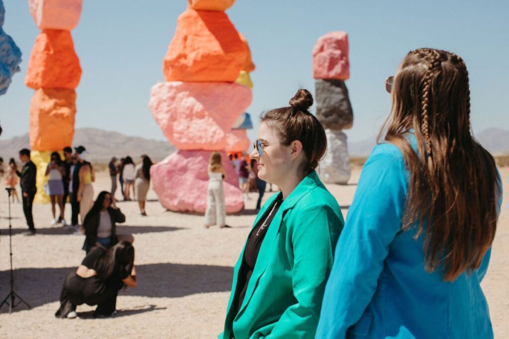 Two women standing together with groups of people behind them standing around colorful rocks. 