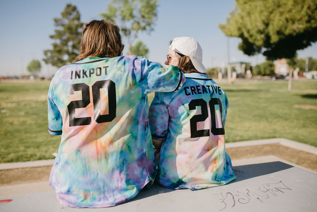 Two women in tie dyed jerseys sitting on a picnic table at a park while one rests their arm on the other's shoulder. 