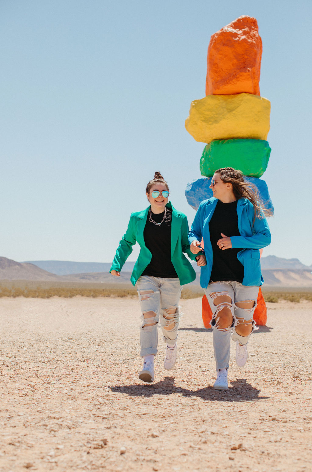 Two women holding hands and running in a desert with a stack of colorful rocks behind them.
