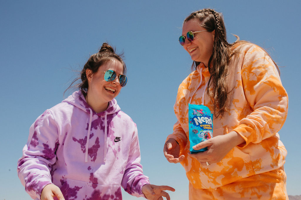 Two women in colorful tie dyed sweatshirts laughing as one holds a bag of candy. 