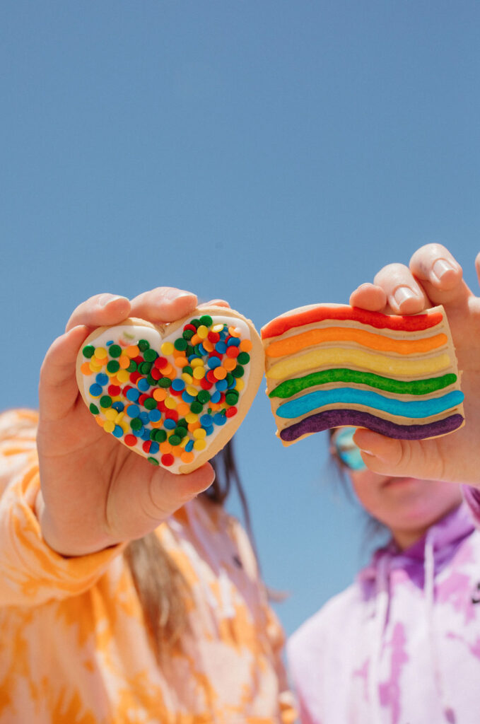 Two women holding out cookies that are decorated with rainbow icing and sprinkles. 