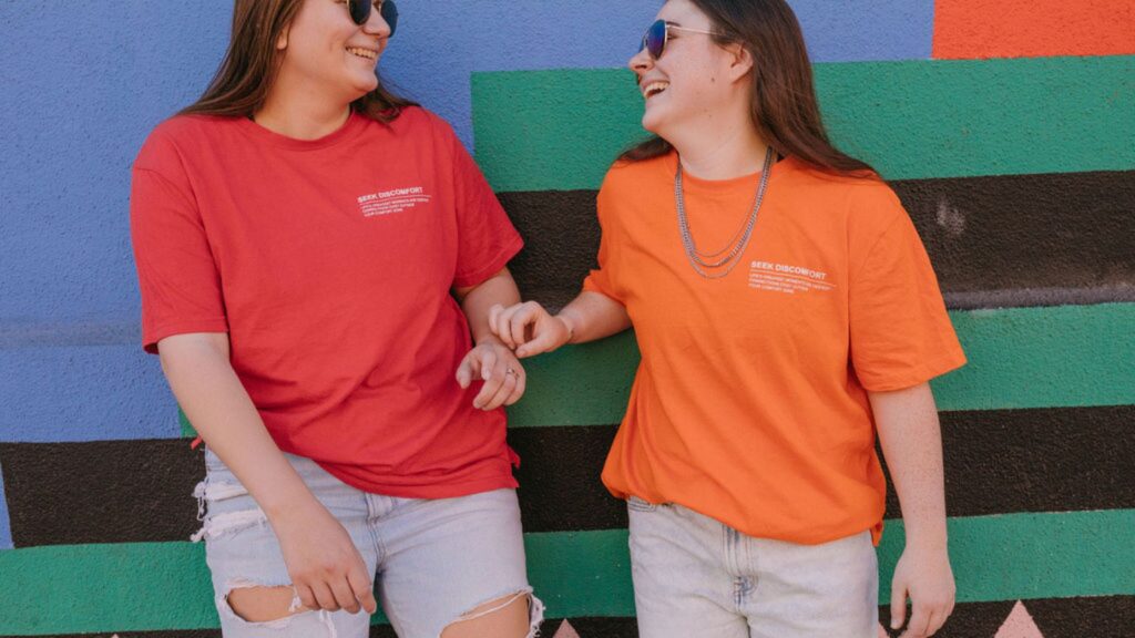 Two women standing against a colorful wall as they laugh and smile together. 
