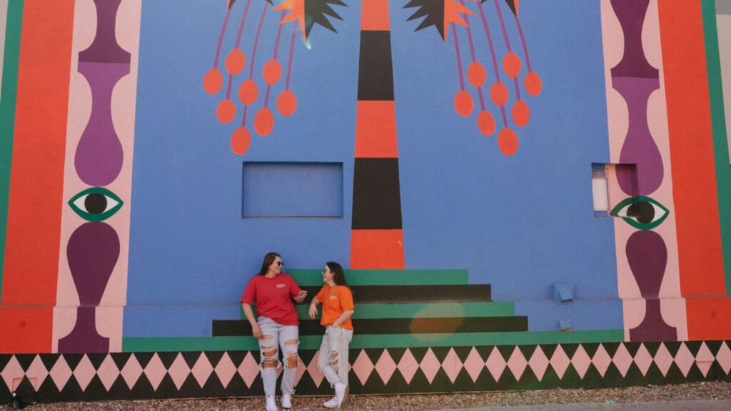 Two women standing against a colorfully painted wall smiling at each other. 
