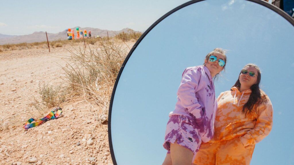 Image of two women in tie dyed sweat suits in the reflection of a circular mirror as they stand in the desert. 

