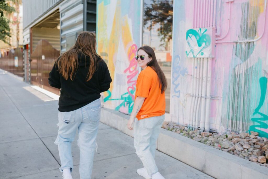 Two girls walking a sidewalk together as one looks back over their shoulder. 