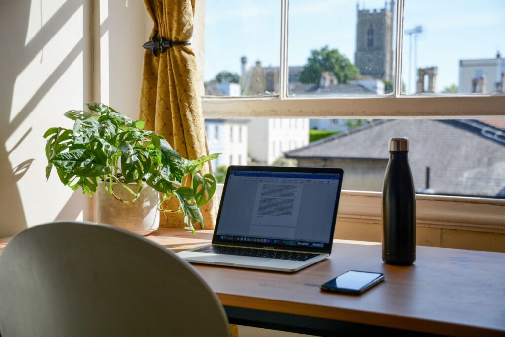 A desk with a laptop open on it in front of a window. 
