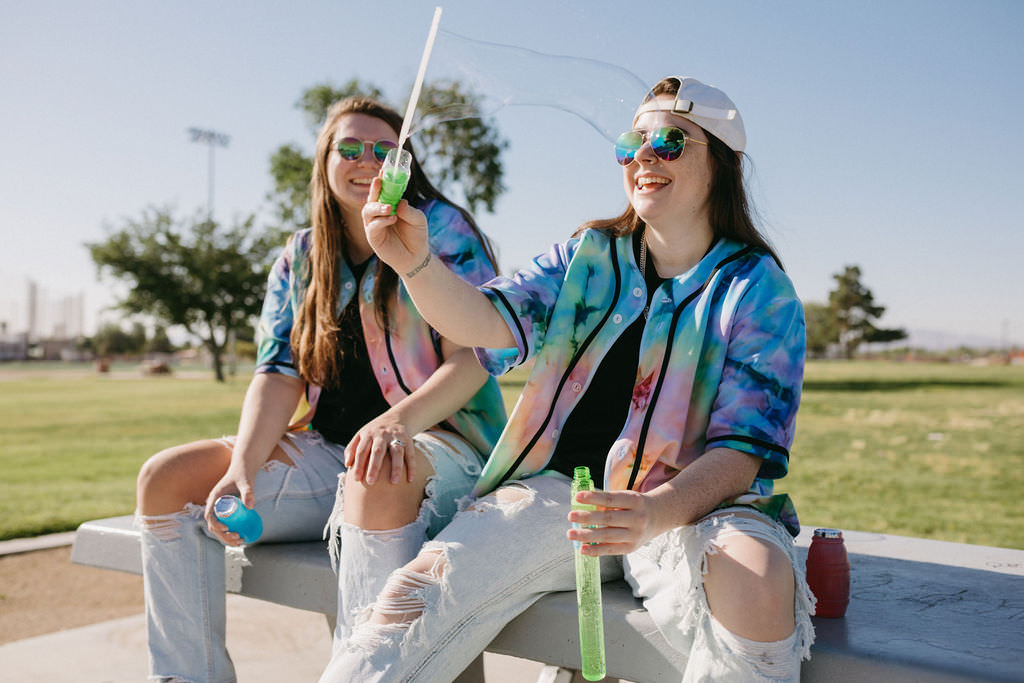 Two people sitting on a picnic table smiling while one of them is blowing bubbles.
