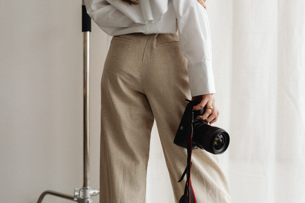 Fashionably dressed photographer with a DSLR camera hanging from her shoulder, standing in a well-lit room.