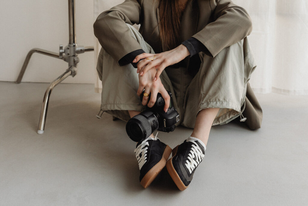 Casual photographer sitting on the floor, holding a DSLR camera, with stylish black and white sneakers crossed.