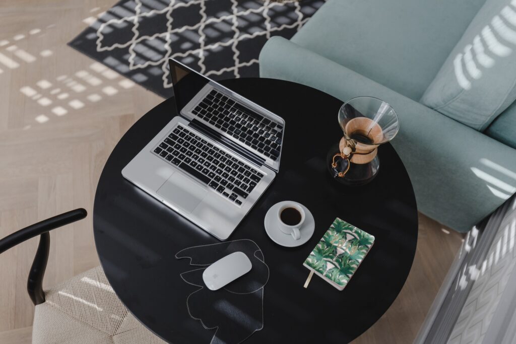 Overhead view of a modern home office with an open MacBook, a coffee cup, a journal with a tropical leaf design, and a pour-over coffee maker on a round black table with a geometric patterned rug and a teal sofa in the background.