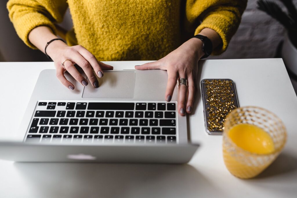 Close-up of a woman's hands using a MacBook on a white table with a gold-sequined smartphone case and a glass of orange juice, representing a vibrant and stylish workspace.