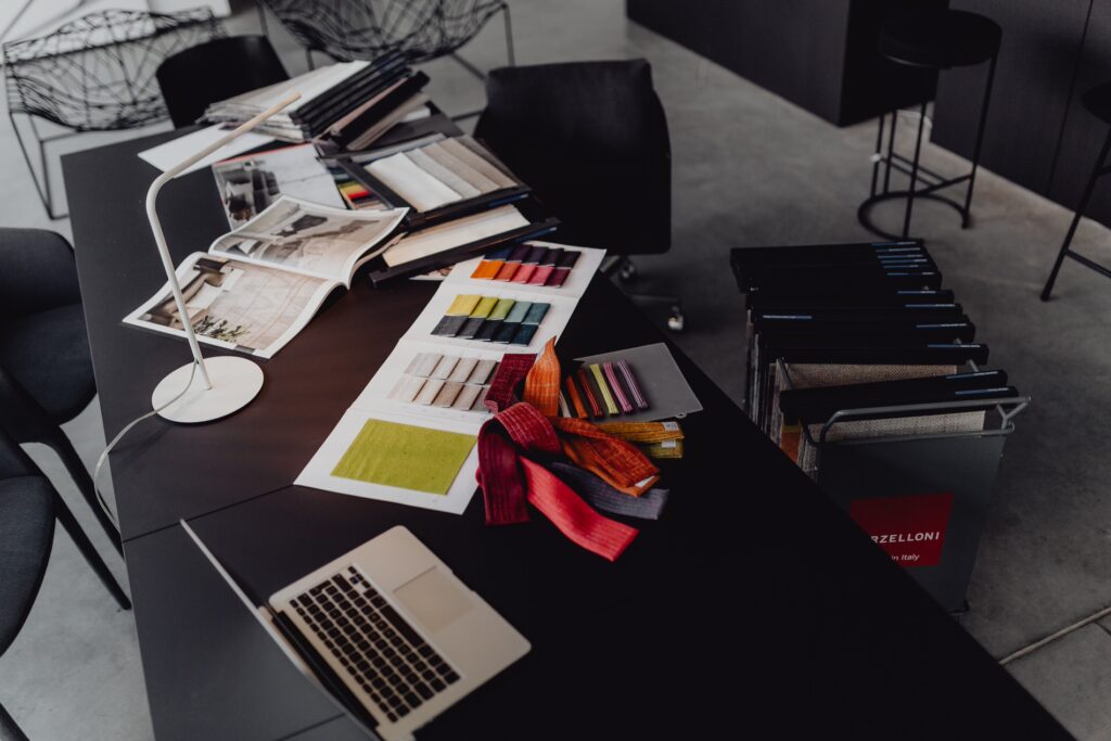 Modern interior design workspace with an assortment of fabric samples spread on a dark table, next to a MacBook and an adjustable white lamp, hinting at material selection for home decor.