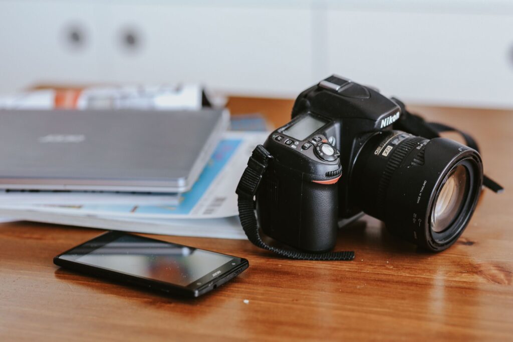Professional Nikon DSLR camera and a smartphone placed on a wooden desk with scattered magazines and a laptop in the background, depicting a photographer's workspace.