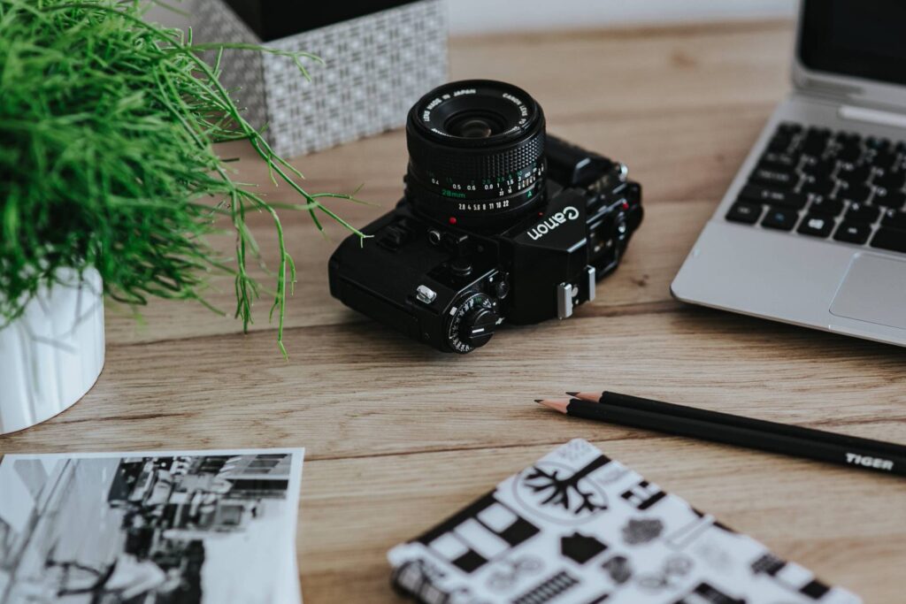 A vintage Canon film camera with a prime lens set beside a MacBook, a black pen, and black and white printed photos, creating a creative workspace atmosphere.
