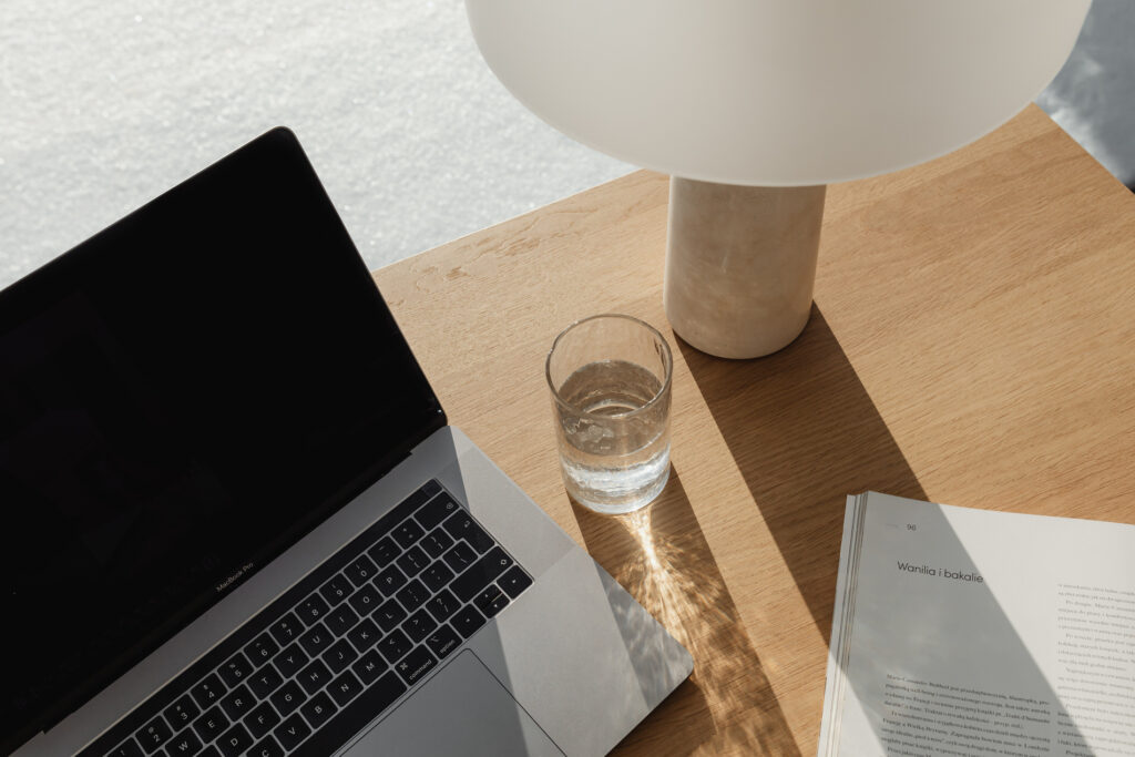 A minimalist office setup with a MacBook Pro, a glass of water, and an open book with visible text "Wanilia i bakalie" under a contemporary lamp, on a wooden table with sunlight casting geometric shadows.