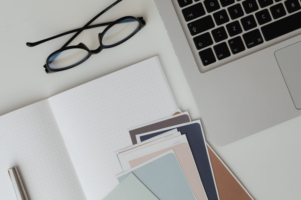 A neat workspace with an open dotted notebook, a pair of black eyeglasses, color swatches, and a MacBook, symbolizing organized creative planning.