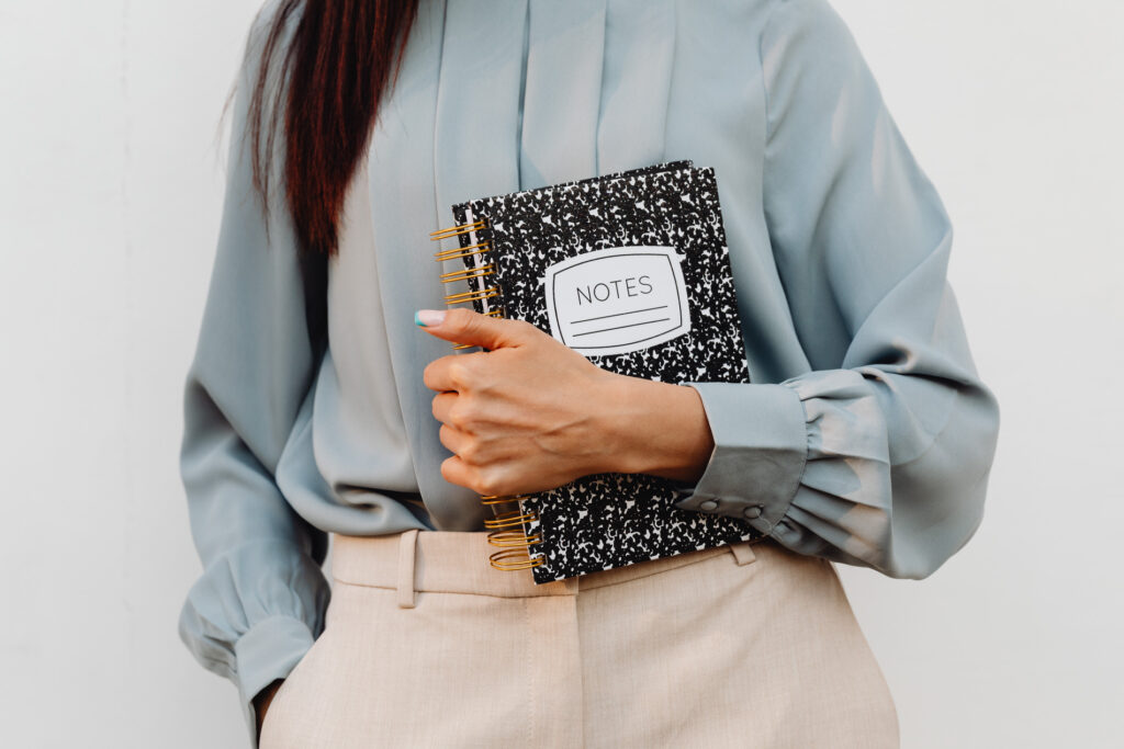 A person in a blue blouse holding a black spiral notebook with 'NOTES' on the cover, suggesting a professional setting for planning or organizing.