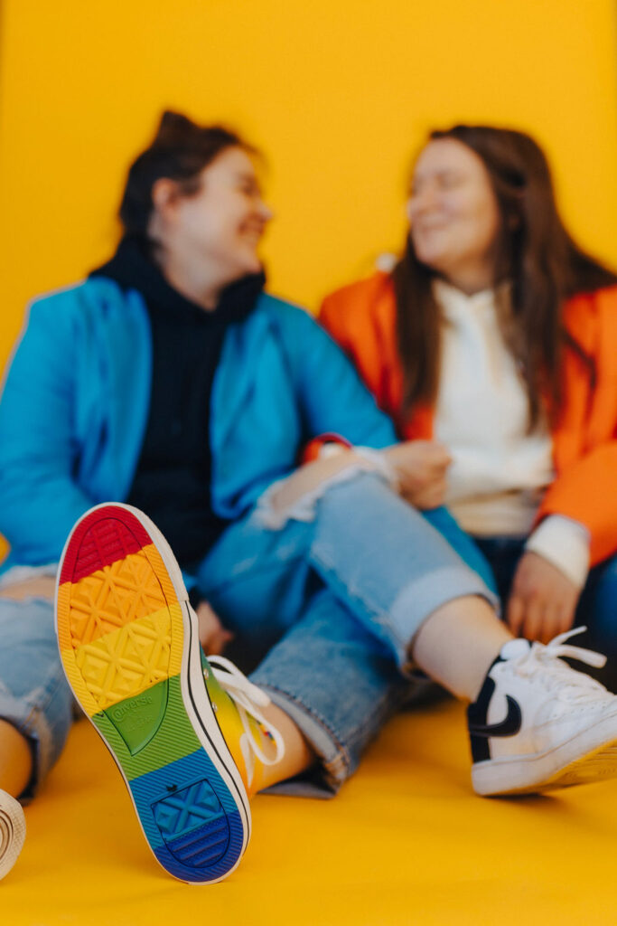 Out-of-focus shot of two women in vibrant clothing, foregrounded by a sneaker with a rainbow-colored sole in sharp detail against a yellow background.