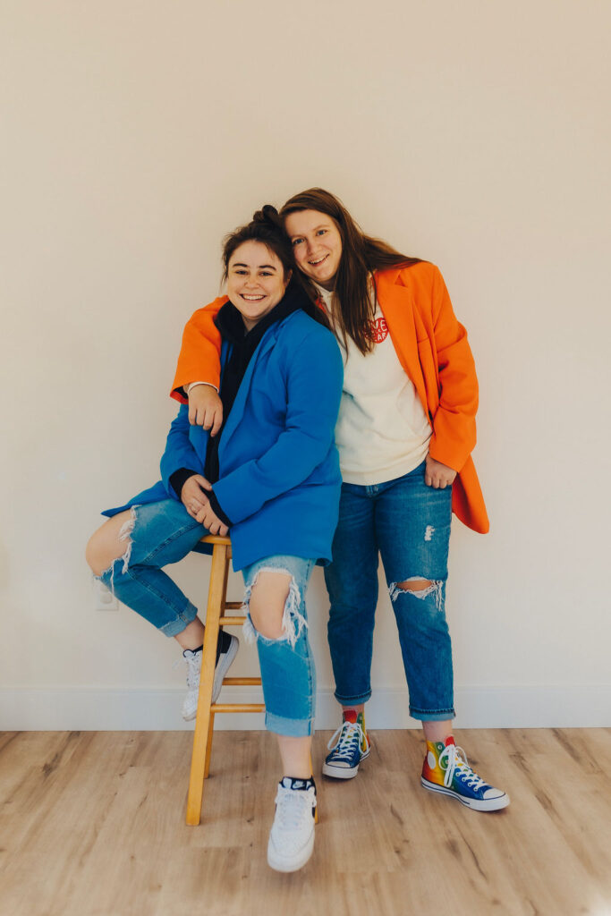 Two women smiling and sitting close together, one on a wooden stool, wearing colorful blazers and distressed jeans in a minimalist setting.