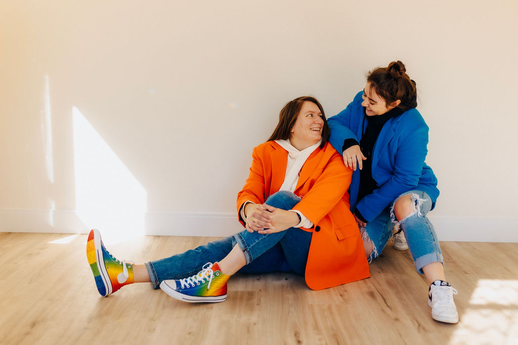 A heartwarming moment between two women sitting on the floor, one wearing an orange blazer and the other in a blue jacket, sharing a joyful interaction.