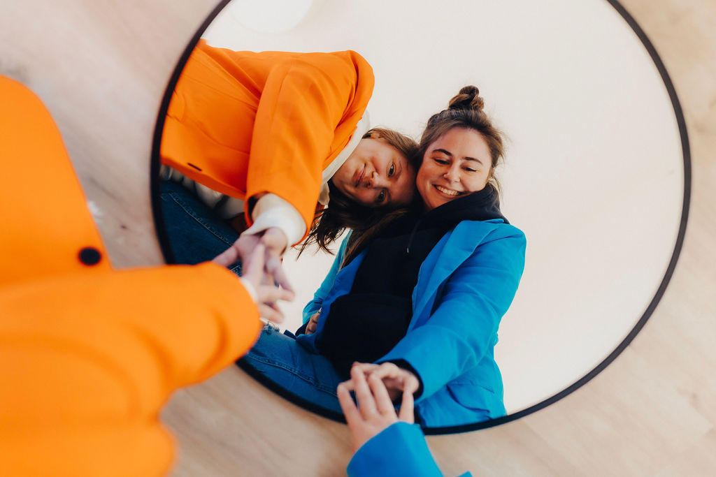 Two women reflected in a round mirror, one in an orange blazer and the other in a blue jacket, both smiling and sitting on the floor.