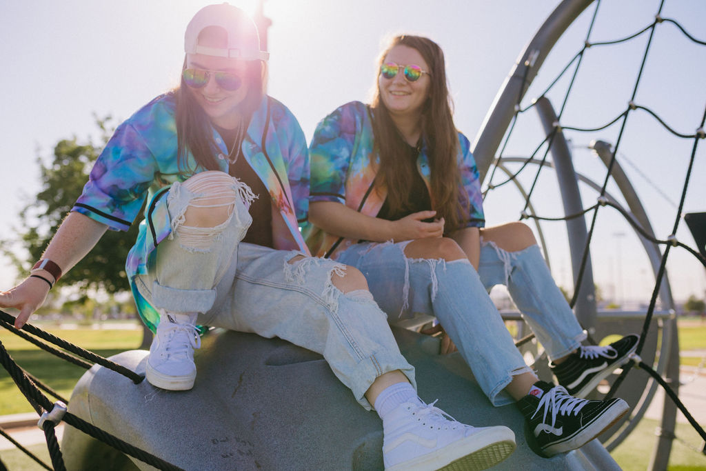Two friends smiling while seated on a playground climbing structure, wearing colorful tie-dye shirts, distressed jeans, and sunglasses.