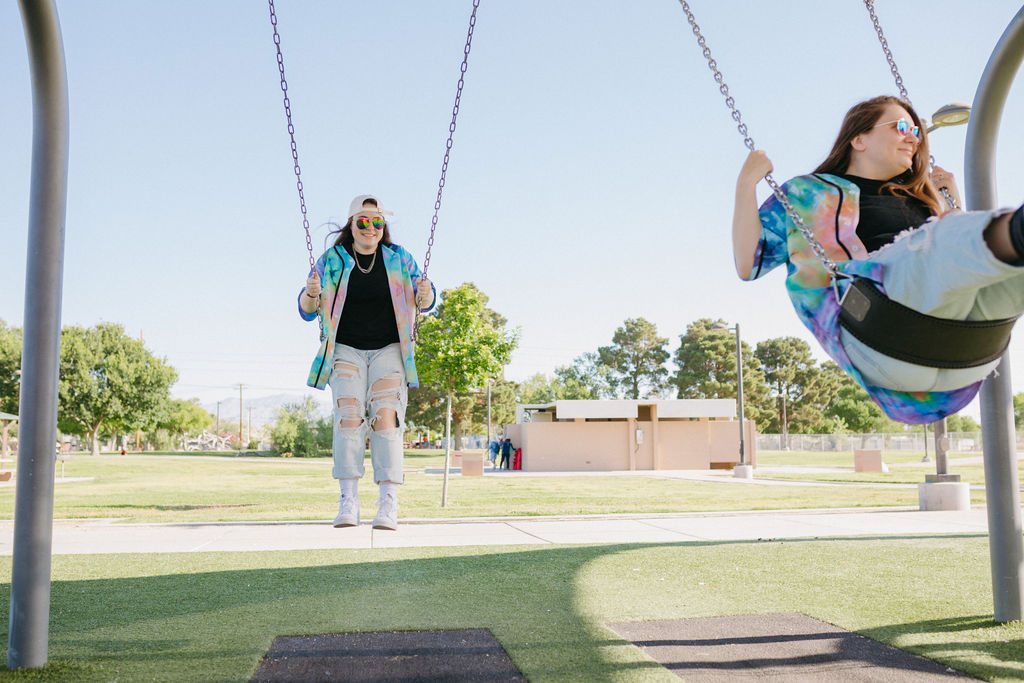A joyful scene at the park with one person swinging high while another sits on a swing, both sporting casual attire and tie-dye shirts.
