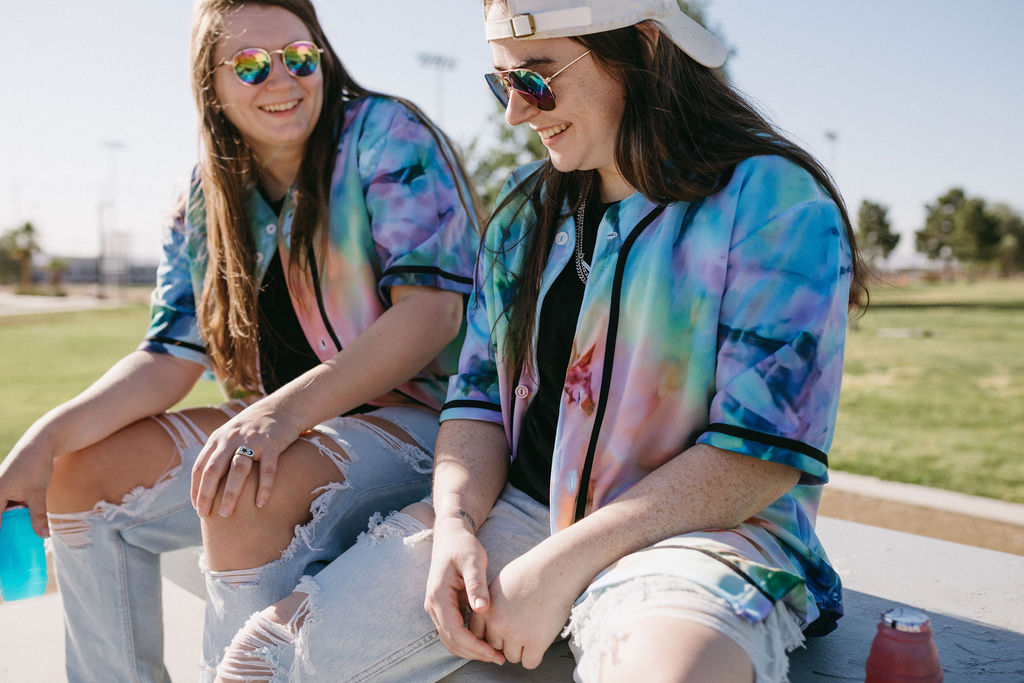 Casual outdoor relaxation captured as two friends sit on a bench, enjoying drinks and conversation, dressed in vibrant tie-dye shirts.
