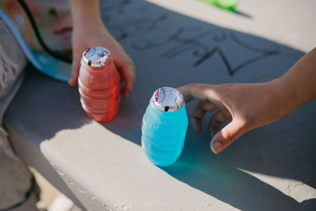 Close-up of two flavored drinks on a concrete surface, with a hand reaching for the blue bottle.