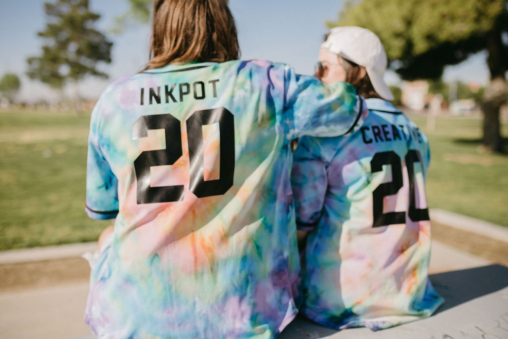 Rear view of two individuals on a park bench, showcasing 'INKPOT 20' and 'CREATIVE 20' on the back of their tie-dye shirts.
