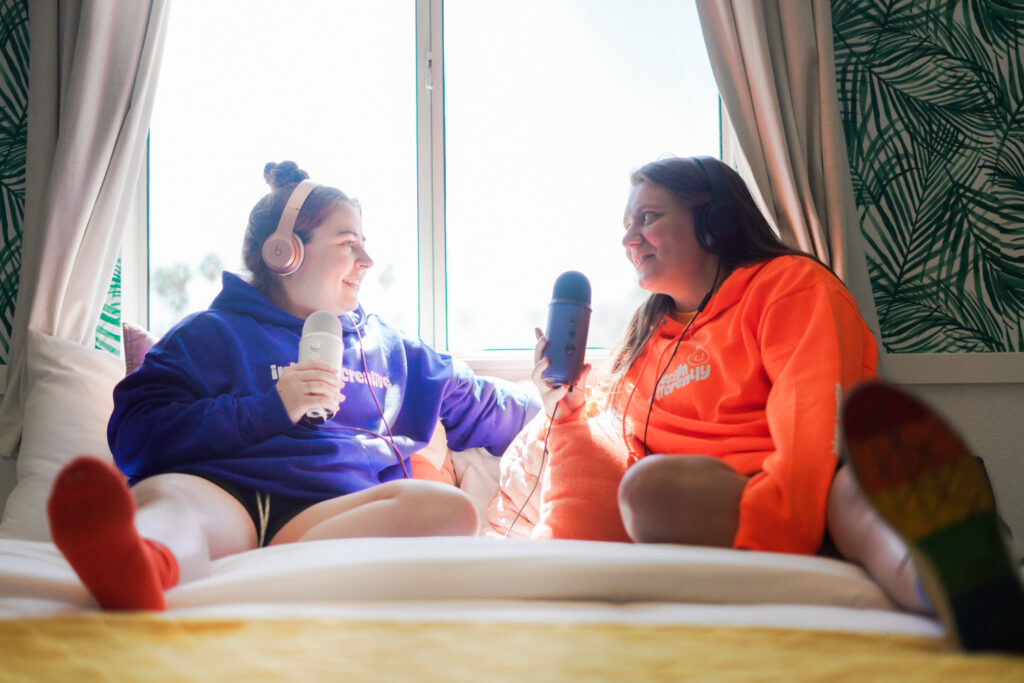 KP and Jessie sit on a bed with podcast mics.