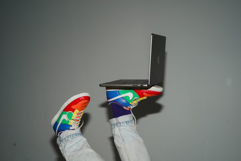 A pair of legs in light-wash jeans and purple socks, wearing colorful sneakers, balancing a laptop on their feet against a gray background. ​​